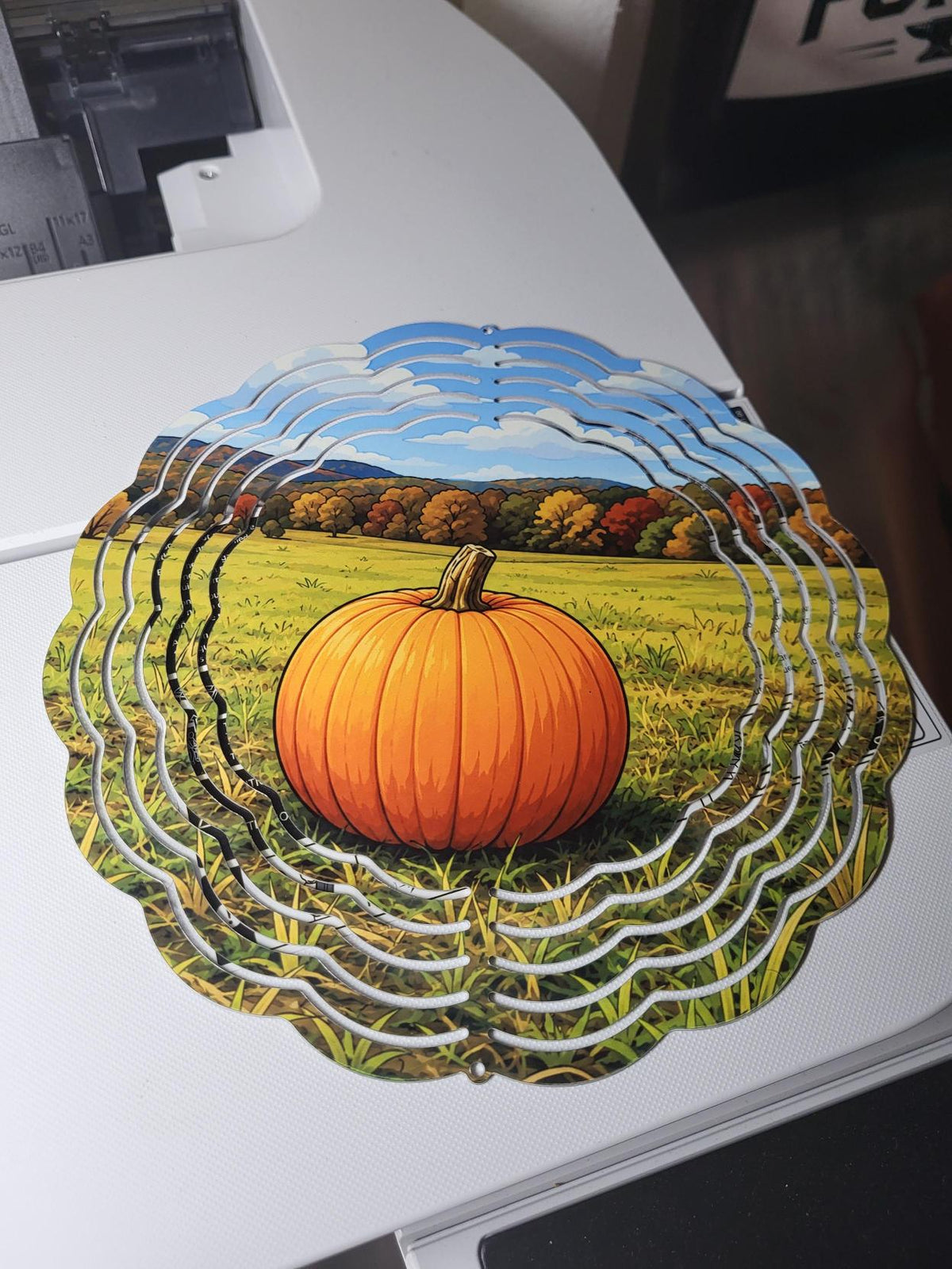 Wind spinner showing a bright orange pumpkin in a sunny autumn field with colorful fall foliage and blue sky