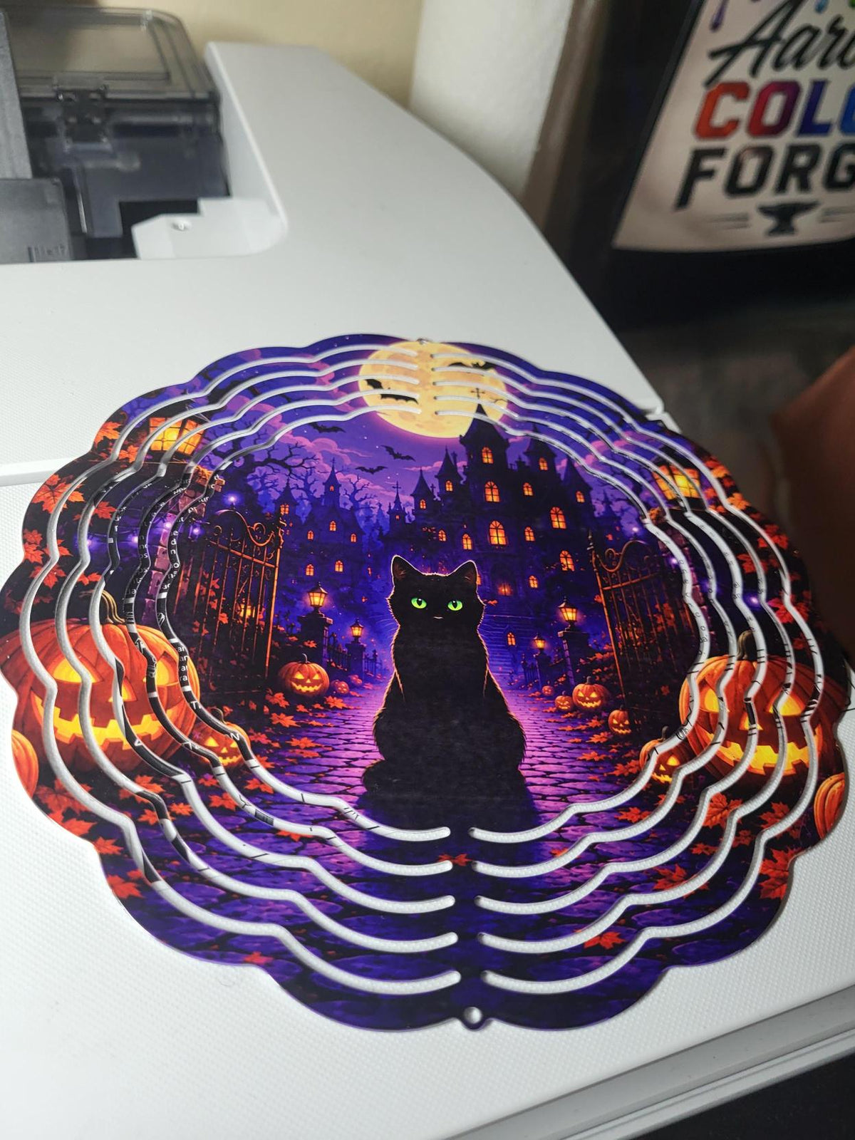 Halloween wind spinner showing black cat with green glowing eyes, iron gate, pumpkins and haunted mansion under full moon